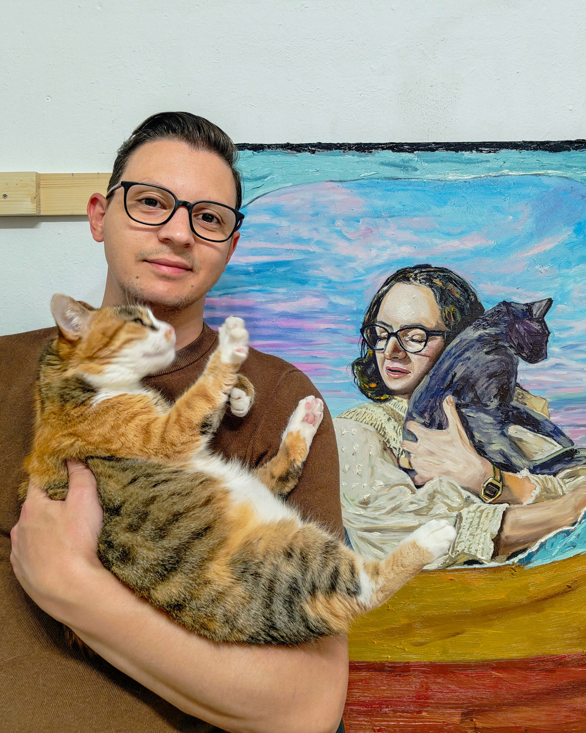 A man wearing glasses and a brown shirt holds an orange and white tabby cat in his arms, standing in front of a colorful painting of a woman holding a black cat.